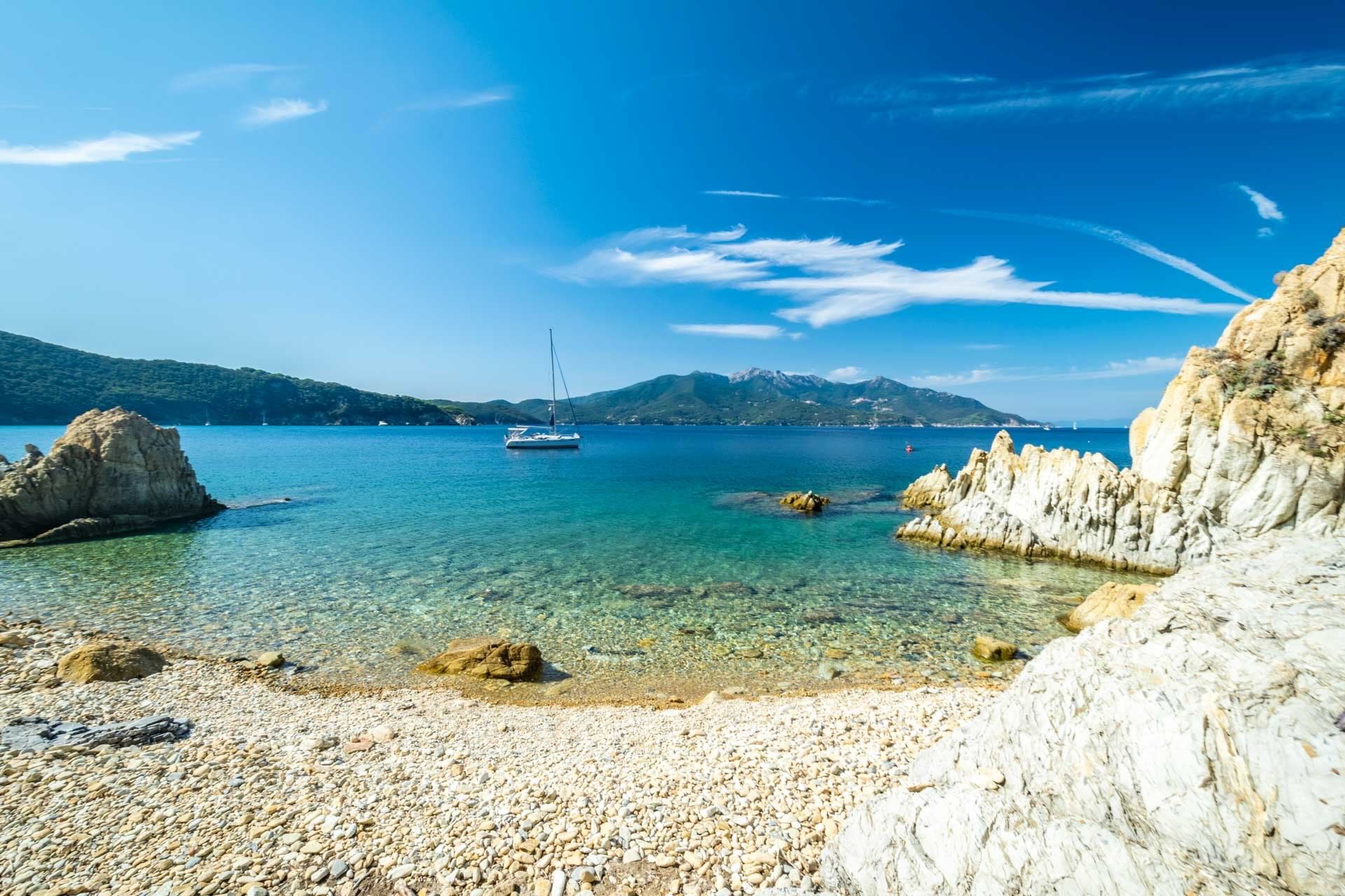 Spiaggia La Pinetina all’Isola d’Elba, con ciottoli bianchi, mare turchese e barca a vela al largo.